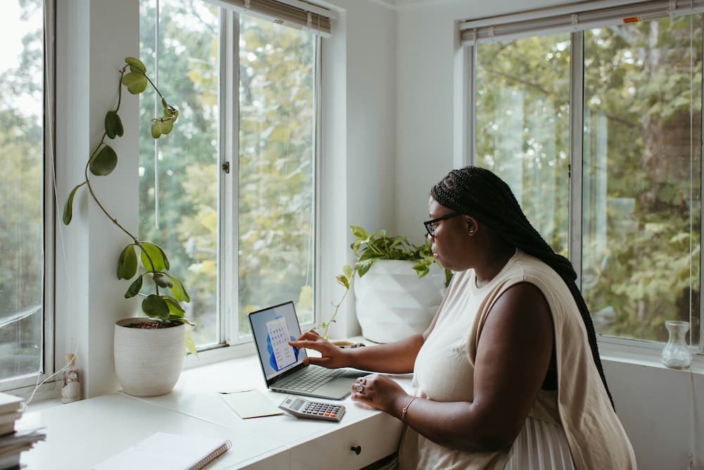 Woman working in a modern home office with a plant by the window, using a laptop and calculator.