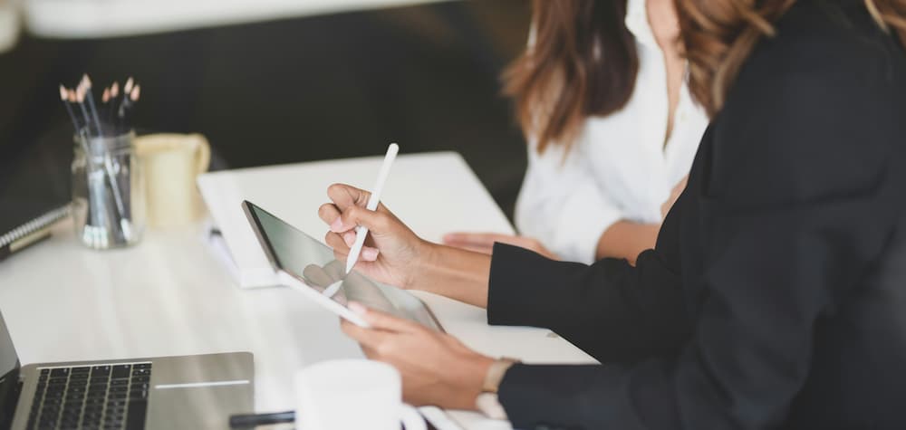 Woman using a stylus on a tablet during a work meeting, with a laptop and notes in the background in an office setting