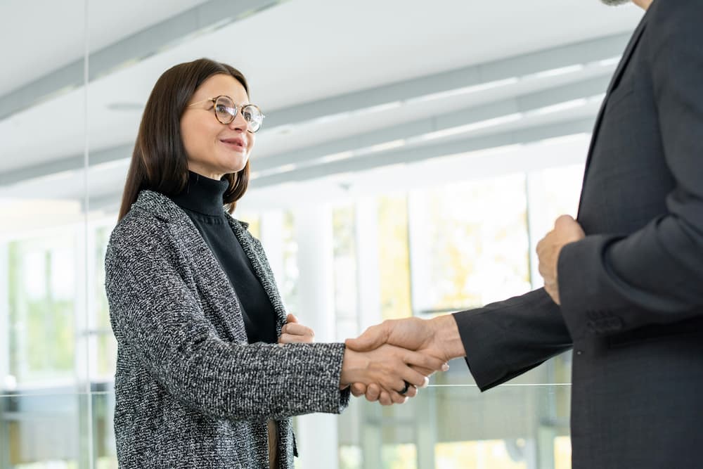 A female lawyer in a grey blazer and black turtleneck is shaking hands with a male client in a dark suit.