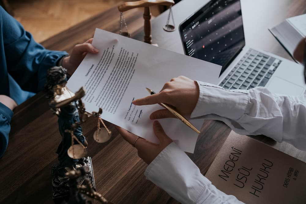 Legal consultation, where a person presents a document to another in an office environment. One person is holding a pen and in the background there is a laptop and a statue of justice.