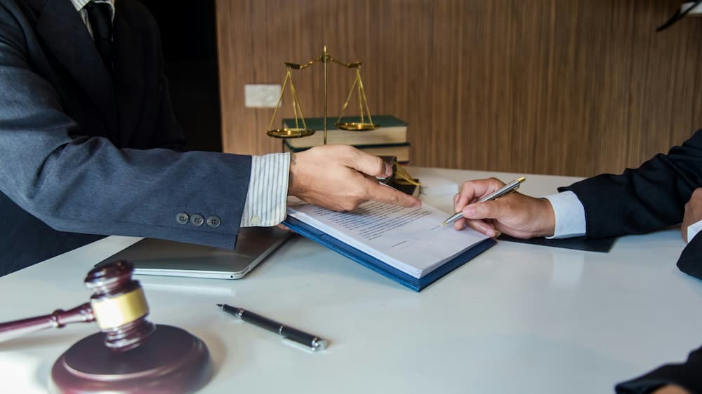 Two individuals in business attire are seated at a desk, engaged in a contract signing. One person is pointing at a document while the other is holding a pen, ready to sign.