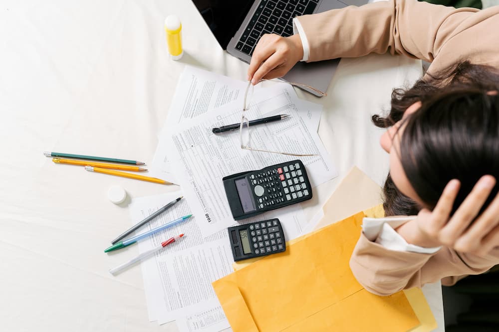 "A person in a beige suit is sitting at a desk cluttered with financial documents, two calculators, various colored pens and pencils, a yellow envelope, and a laptop.