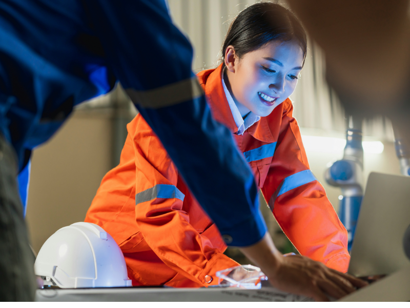 A female engineer or technician in an orange safety uniform is smiling and engaged while reviewing information on a laptop.
