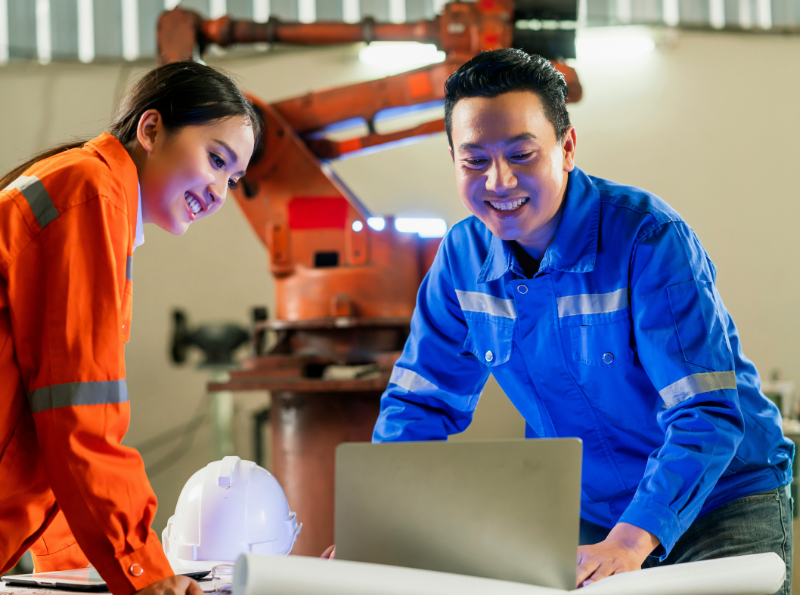 A male and a female engineer are smiling and collaborating while working on a laptop in a manufacturing environment.