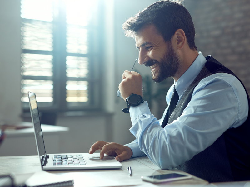 The image shows a smiling man sitting at a desk and working on a laptop. He's dressed in a light-blue shirt and a dark vest, with a watch on his wrist. With one hand, he's holding a pair of glasses, and with the other, he's using the laptop's touchpad.