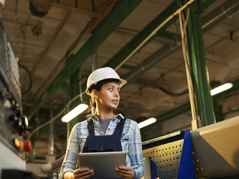 A female worker or engineer is seen standing in an industrial or manufacturing facility. She is wearing a white safety helmet and a blue denim shirt with overalls, indicating that she is likely involved in a technical or supervisory role on the factory floor.