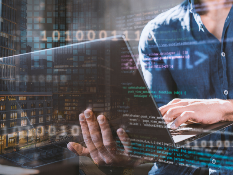 
This image is a double exposure that combines two separate scenes.

In the foreground, a person wearing a blue shirt is holding and typing on a laptop. The screen of the laptop is filled with lines of computer code.